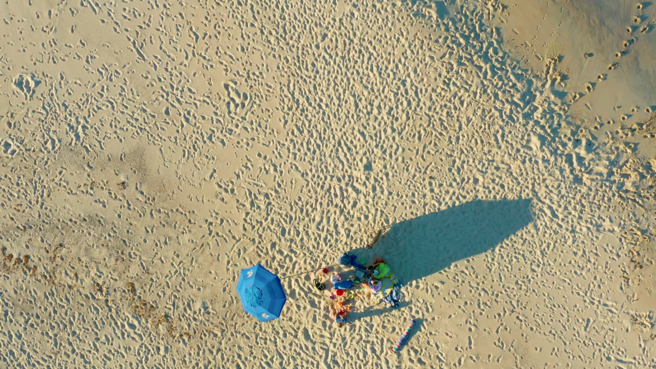 Top-down view of people relaxing on a sandy Las Barracas beach near a blue umbrella, with footprints and elongated shadows visible in the afternoon-like light.