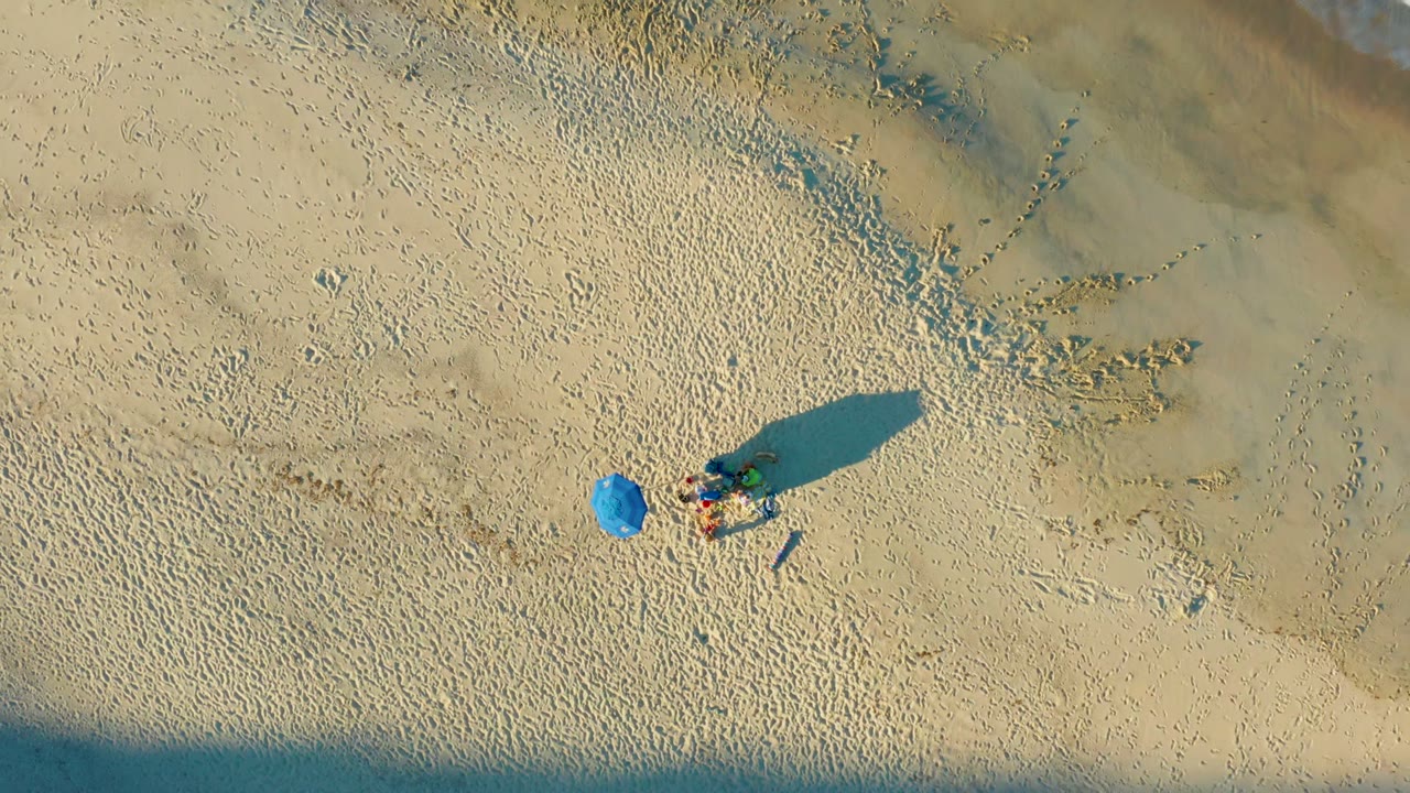 Overhead frame from Las Barracas, Baja California Sur, Mexico, showing a blue umbrella beside a small group of visible people on pale sand with long shadows.