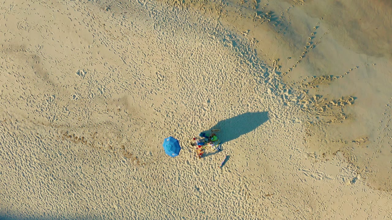 Aerial frame from Las Barracas showing visible beachgoers beside a bright blue umbrella, with open footprint-marked sand and a large shadow extending to the right.