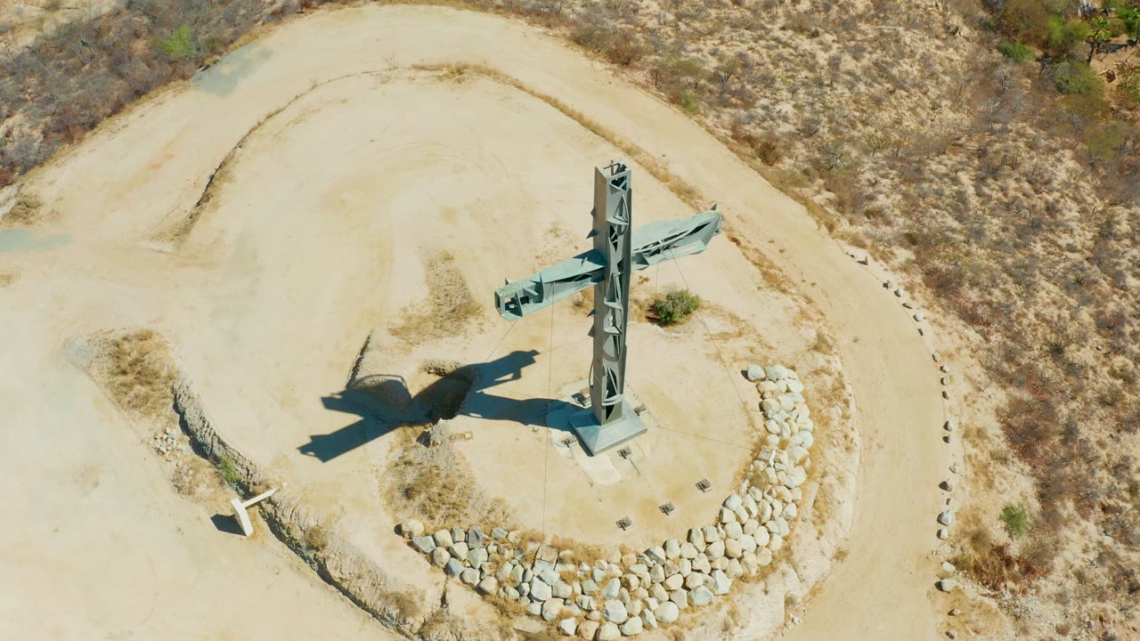 Overhead frame of a tall cross monument on sandy hilltop terrain in Puerto Los Cabos, Baja California Sur, with stone edging below it and no visible people.