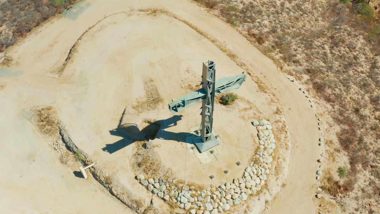 High overhead view of the Puerto Los Cabos cross with its shadow angled across pale sandy ground, scrubland at the top edge, and no visible people.