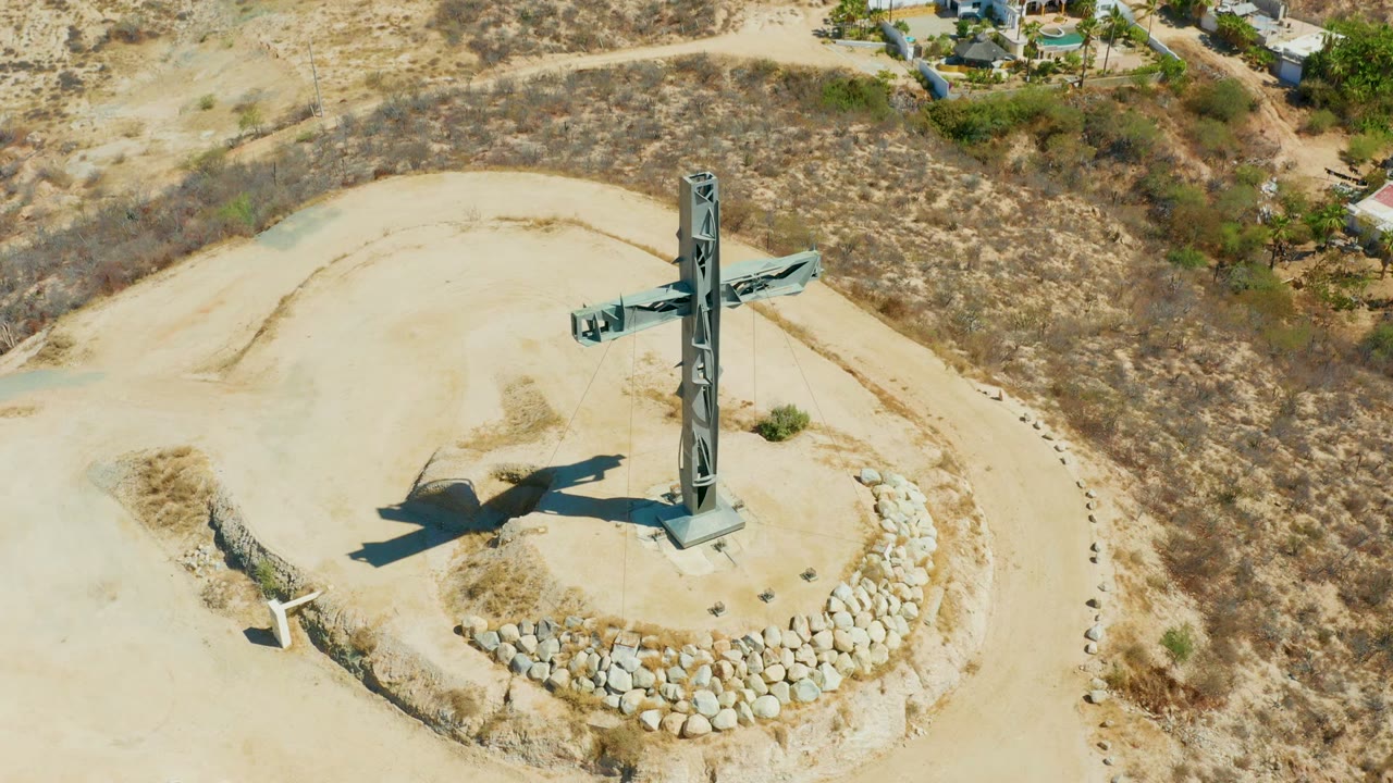 Overhead drone frame of a tall cross monument on a sandy hilltop in Puerto Los Cabos, with a circular stone border, dry brush, and no visible people.