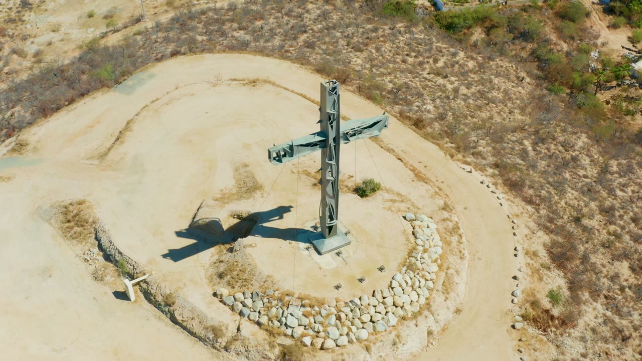 Drone frame over the Puerto Los Cabos cross monument with dry scrubland surrounding the sandy lookout and the circular stone edge visible below, with no visible people.
