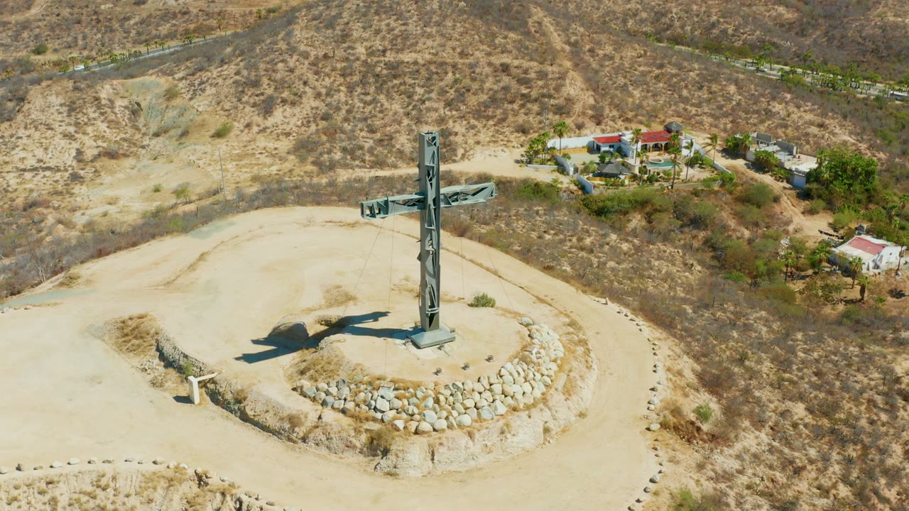 Aerial frame over Puerto Los Cabos showing a tall cross set in a stone circle on a pale dry hilltop, with scrubby slopes, small buildings, a road, and no visible people.