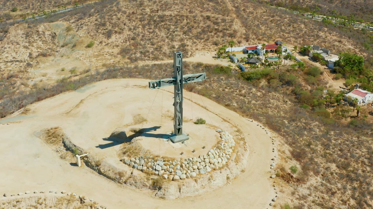 Large metal cross on a hilltop with a long shadow and buildings beyond.