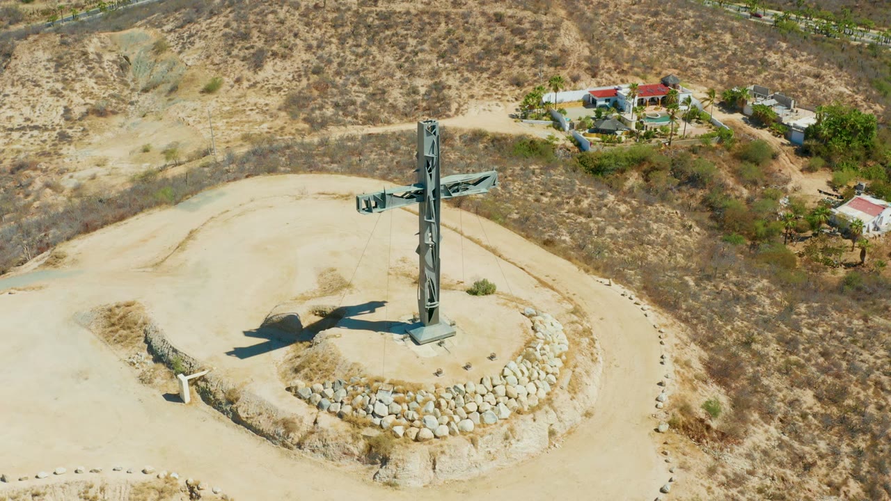 Aerial frame in Puerto Los Cabos showing the hilltop cross upright on its square base, the circular stone border below it, dry brush on the slopes, background buildings, and no visible people.
