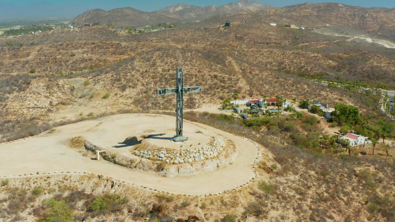 Aerial view of a tall hilltop cross in Puerto Los Cabos, Baja California Sur, with a circular stone path, dry scrubland, nearby buildings, distant mountains, and no visible people.
