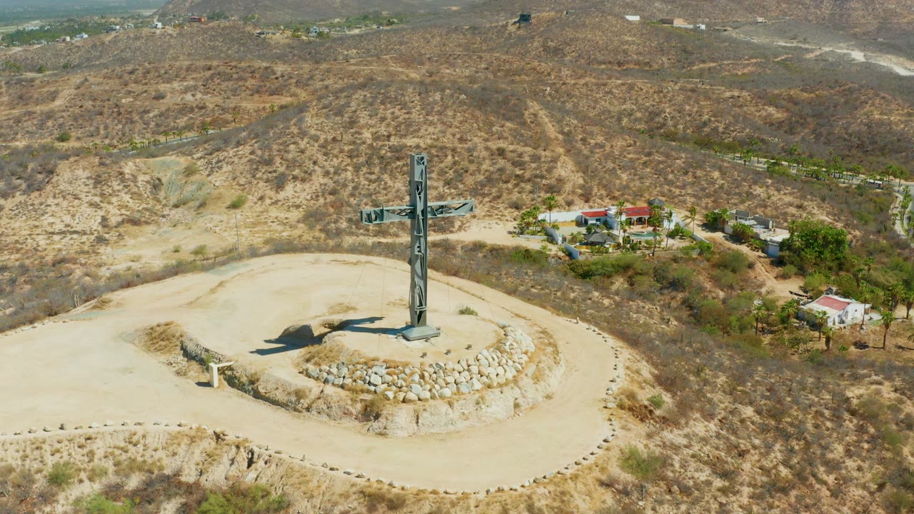 A centered aerial composition shows the hilltop cross in Puerto Los Cabos above a pale circular path, with rocky dry ground, scattered shrubs, buildings at right, and no visible people.