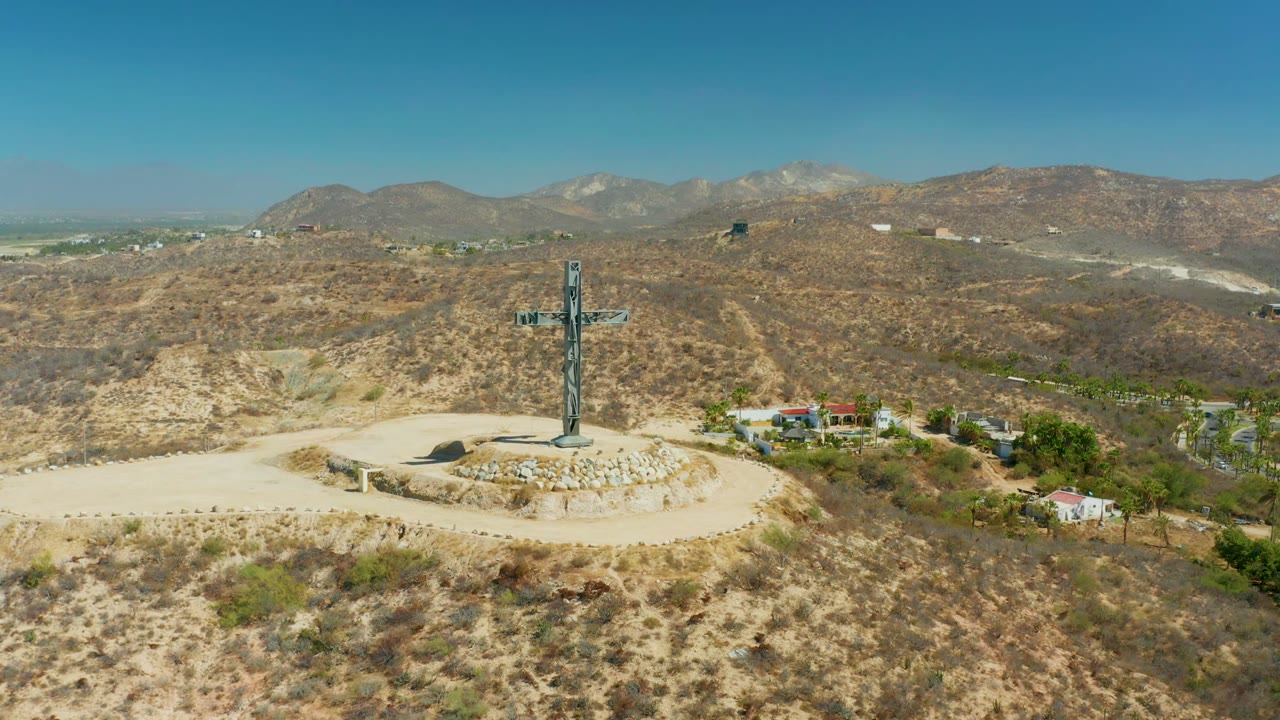 Aerial view of a hilltop cross in Puerto Los Cabos, Baja California Sur, with a circular stone turnout, scrubby dry hills, distant mountains, clear blue sky, and no visible people.