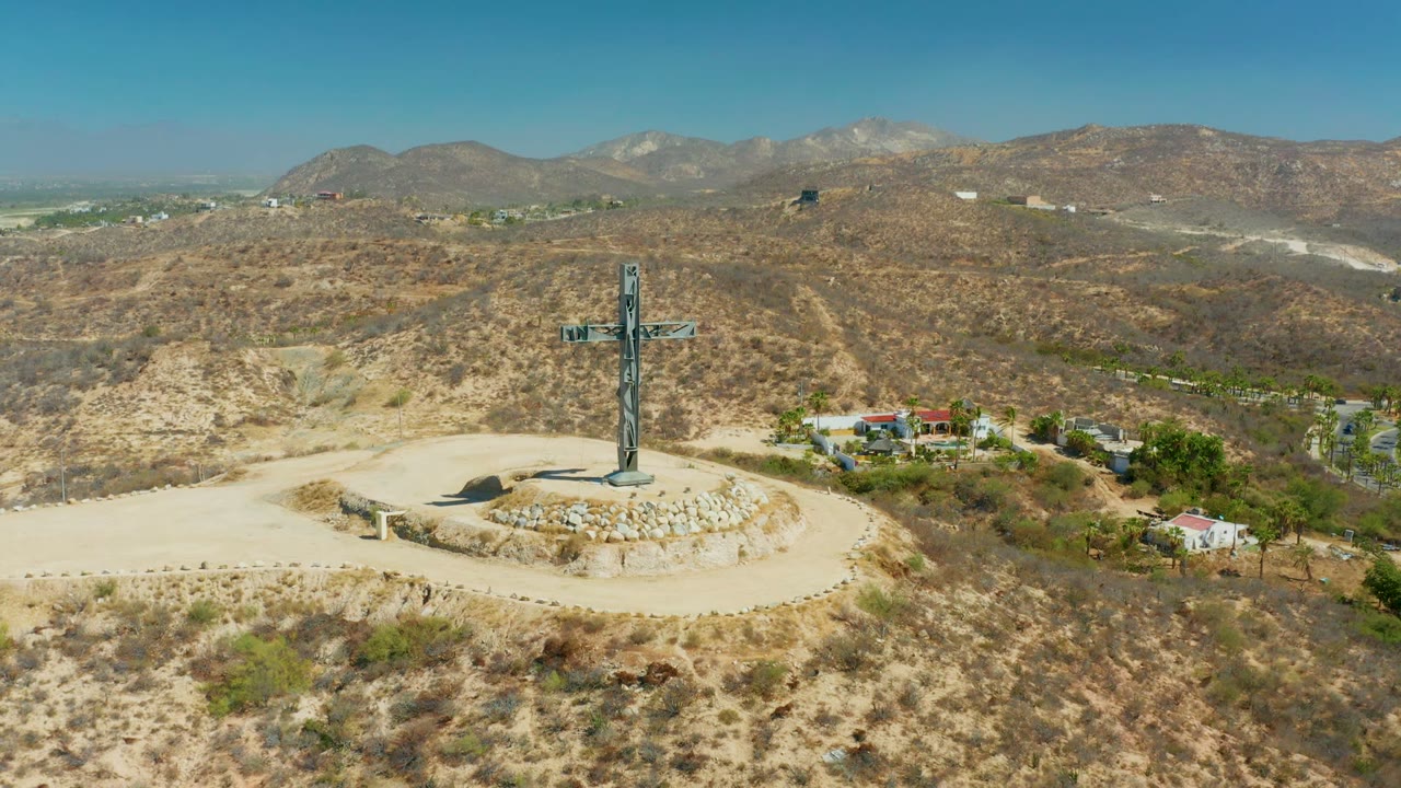 Drone view over Puerto Los Cabos with the hilltop cross on a circular lookout, dry mountains across the background, a small cluster of homes and trees to the right, and no visible people.