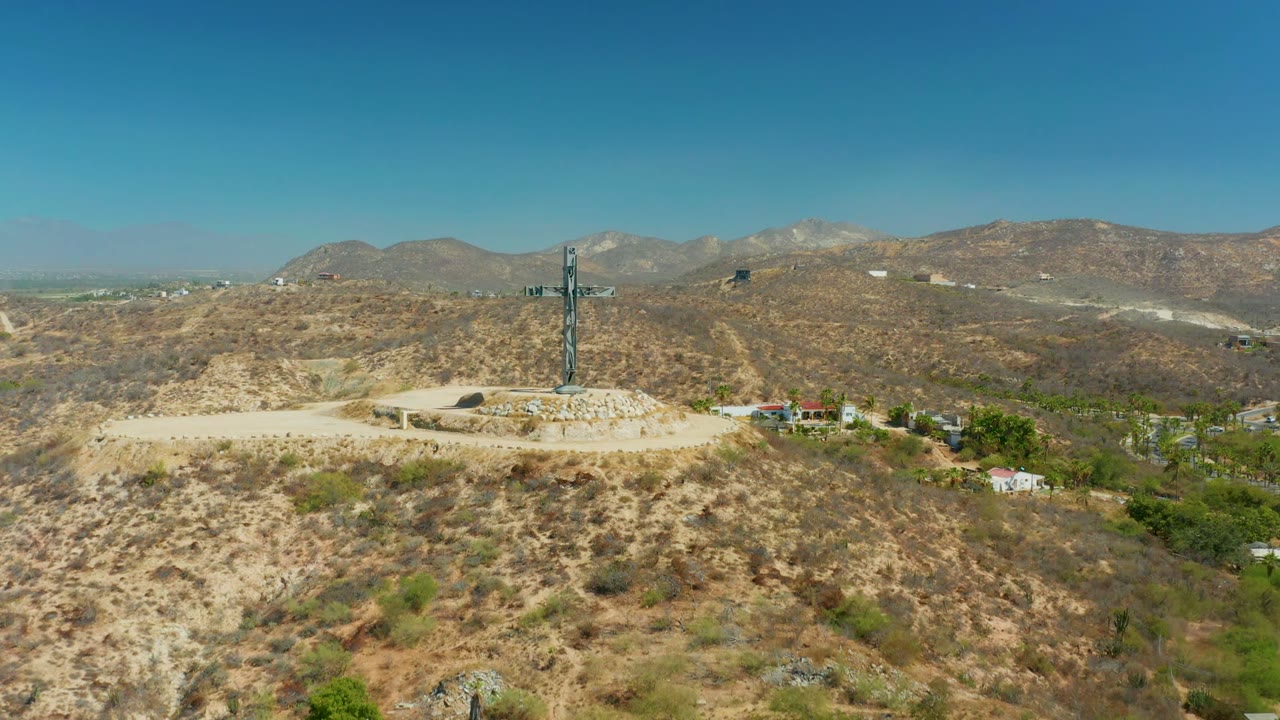 Opening aerial frame of a tall cross on a stone platform in Puerto Los Cabos, with scrubby tan hills, a dirt road, distant mountains, blue sky, and no visible people.