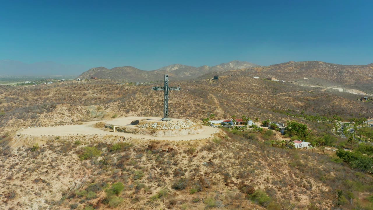 The cross stands on its round stone base in Puerto Los Cabos, with dry foreground brush, small buildings and greener vegetation to the right, mountains behind, and no visible people.
