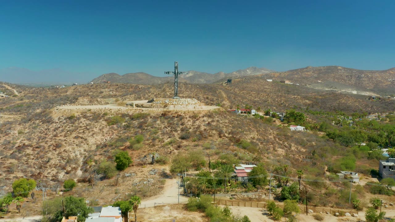 Wide aerial view in Puerto Los Cabos showing a hilltop cross on a stone circle, dry scrub hills, scattered buildings, green trees, distant mountains, and no visible people.