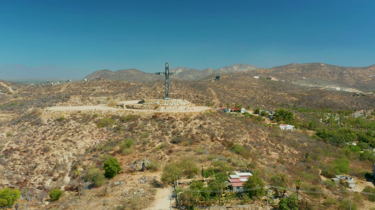 Drone view in Puerto Los Cabos with the hilltop cross near center, dry brush in the foreground, tree clusters and roofs lower in frame, hazy distant mountains, and no visible people.