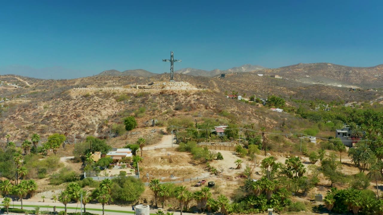 Opening frame of Puerto Los Cabos with a tall hilltop cross, arid slopes, palm trees, small buildings, distant mountains, and no visible people.