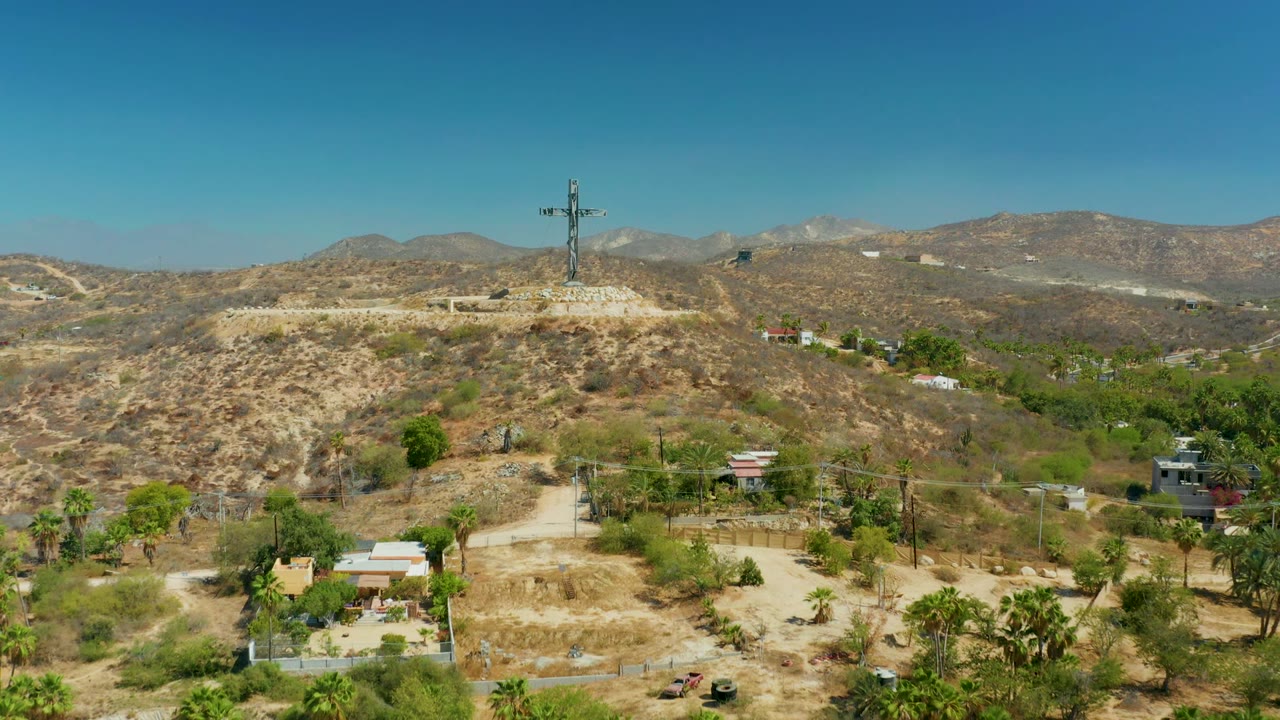 Third frame of the Puerto Los Cabos aerial holds the cross on the hilltop above tan scrub, a few small buildings, palms, and a clear mountain backdrop with no visible people.
