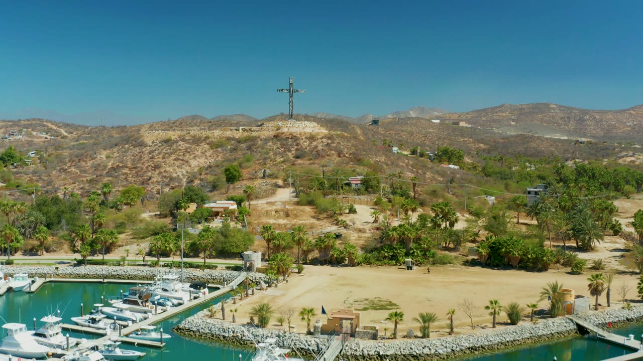 Wide aerial frame in Puerto Los Cabos showing white boats in turquoise marina water, sandy ground, palms, dry hills, distant mountains, and a cross on the ridge, with no visible people.