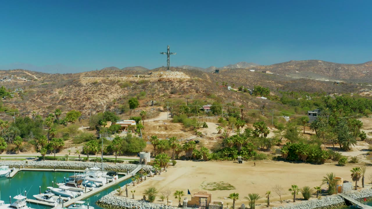 Sandy marina edge and palms beneath a cross on a dry hill.
