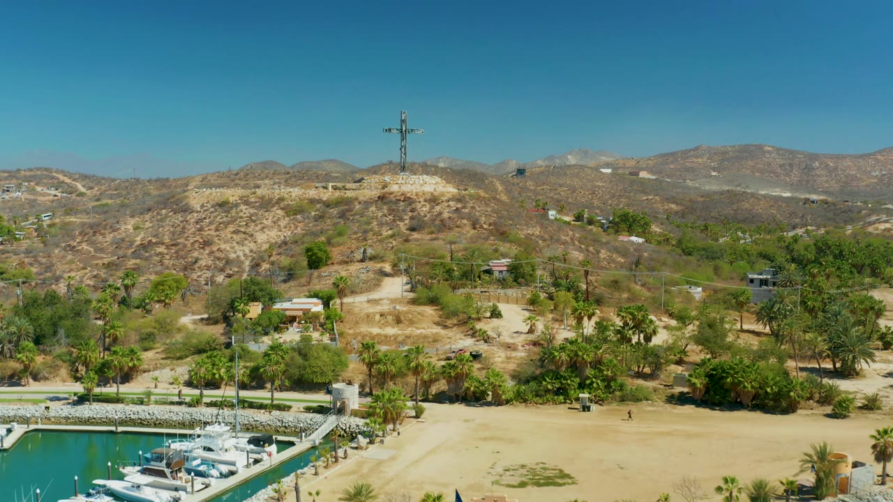 Aerial frame over Puerto Los Cabos with the marina corner at lower left, palms and sandy land across the middle, and a cross on a dry ridge backed by mountains, with no visible people.