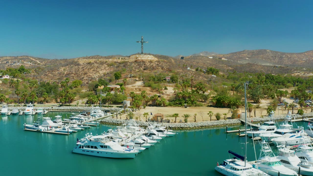 Opening frame of Puerto Los Cabos marina with docked white yachts in blue-green water, rocky shoreline, palms, dry hills, a hilltop cross, clear sky, and no visible people.