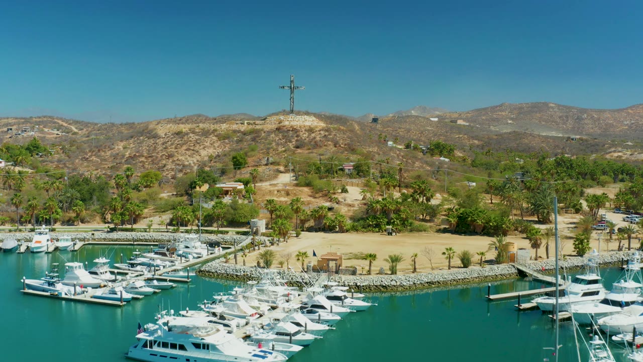 Puerto Los Cabos marina frame with white yachts and boats filling the lower view, dock piers crossing the water, palms along shore, dry hills in back, a hilltop cross, and no visible people.