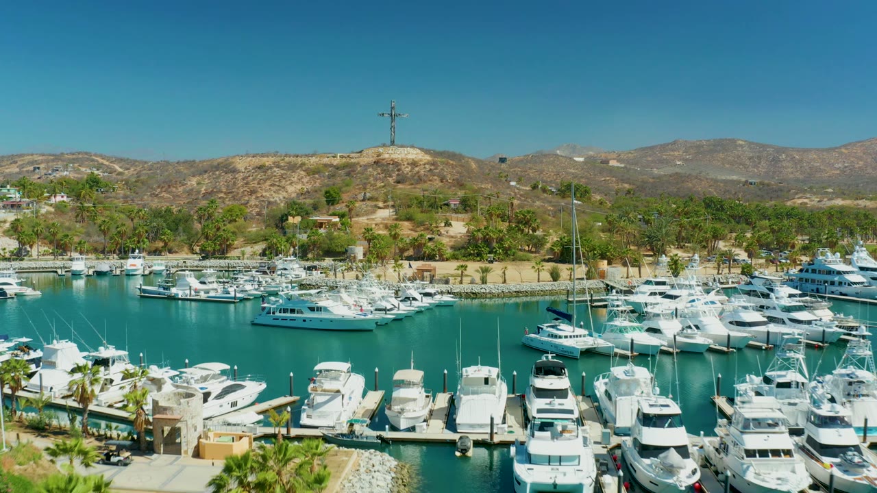 Wide aerial frame of Puerto Los Cabos marina with white yachts in foreground slips, turquoise water, palms, dry hills, a hilltop cross, and no visible people.