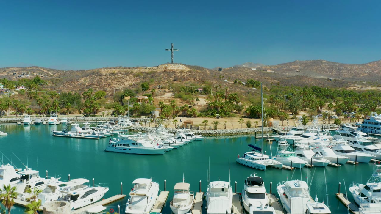 Aerial view over Puerto Los Cabos marina with rows of white yachts in bright sun, teal water, palm-lined shore, hillside terrain, and no visible people.