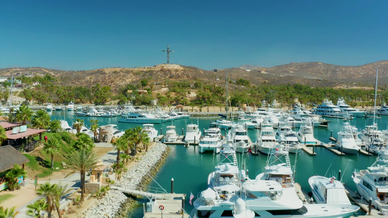 Opening aerial frame of Puerto Los Cabos marina with white yachts in the foreground slips, turquoise water, palms along the shore, dry hills beyond, and no visible people.