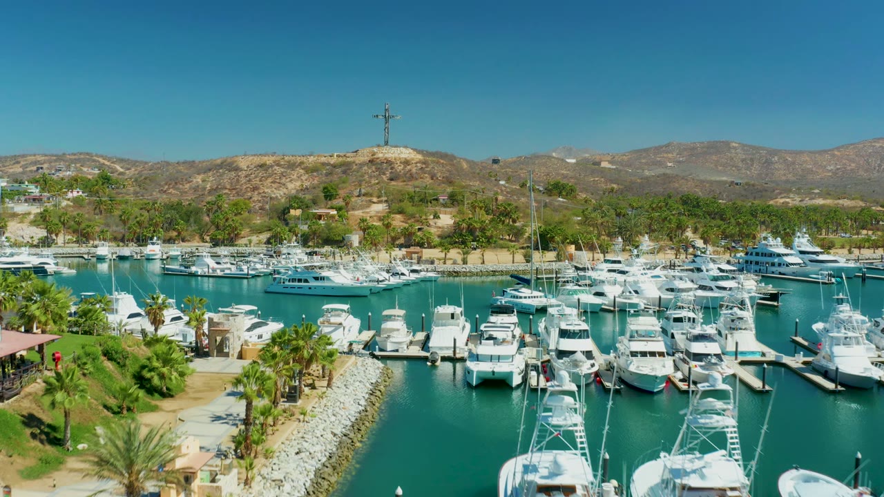 Aerial frame of Puerto Los Cabos marina with docked boats across the water, a rock breakwater in the lower center, palms, dry hills, a hilltop cross structure, and no visible people.