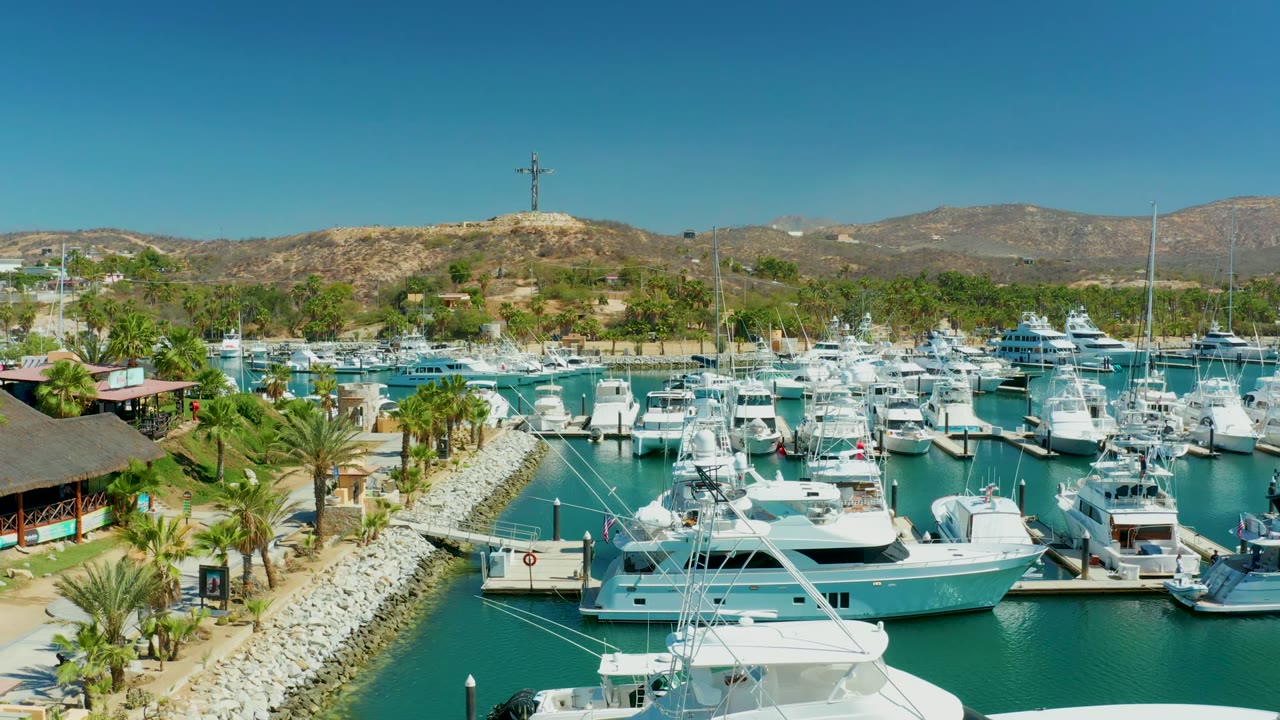 Docked yachts in turquoise marina water with palms, rocky shore, and hills under blue sky.