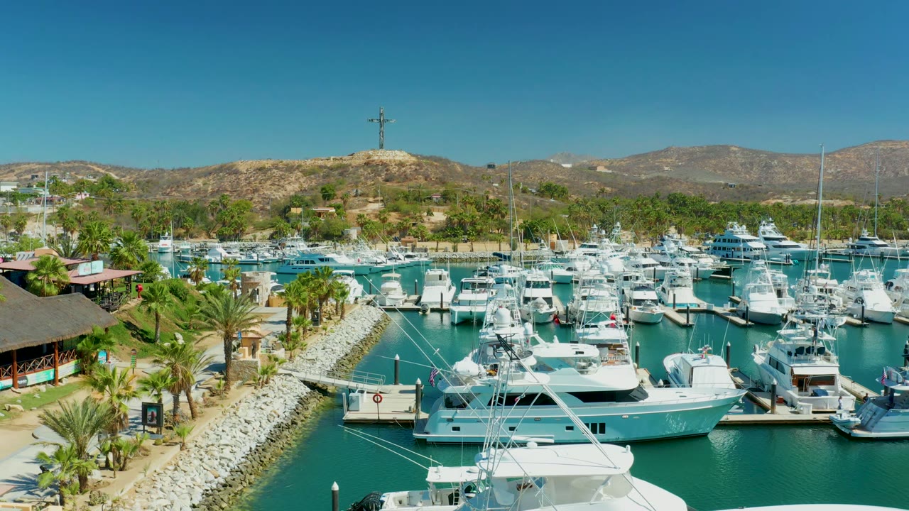 Elevated Puerto Los Cabos marina view with closely packed white yachts, green-blue water, palms and rocks at left, distant hills, clear sky, and no visible people.