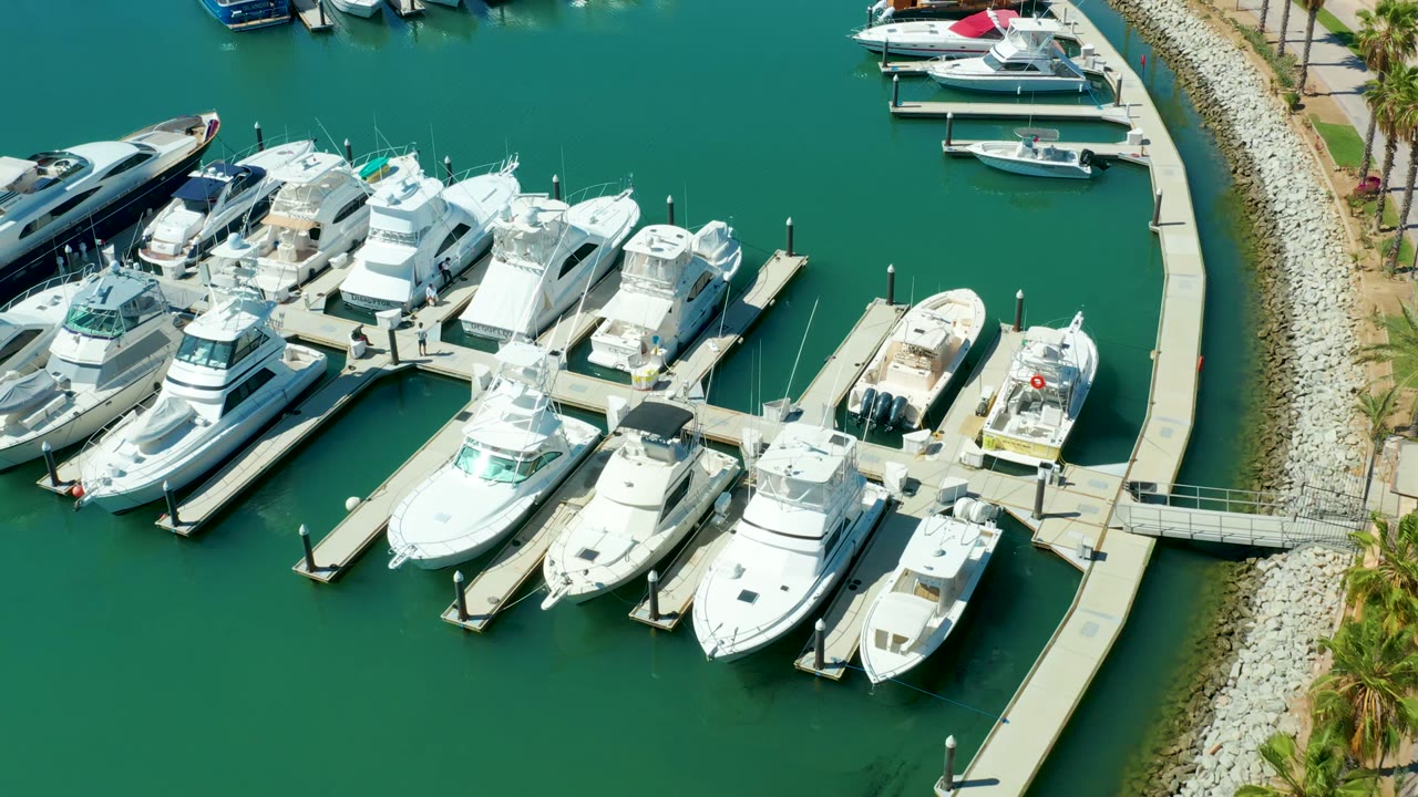 Overhead view of Puerto Los Cabos marina with white yachts packed along docks, a curved pier on the right, green water around the slips, and no visible people.