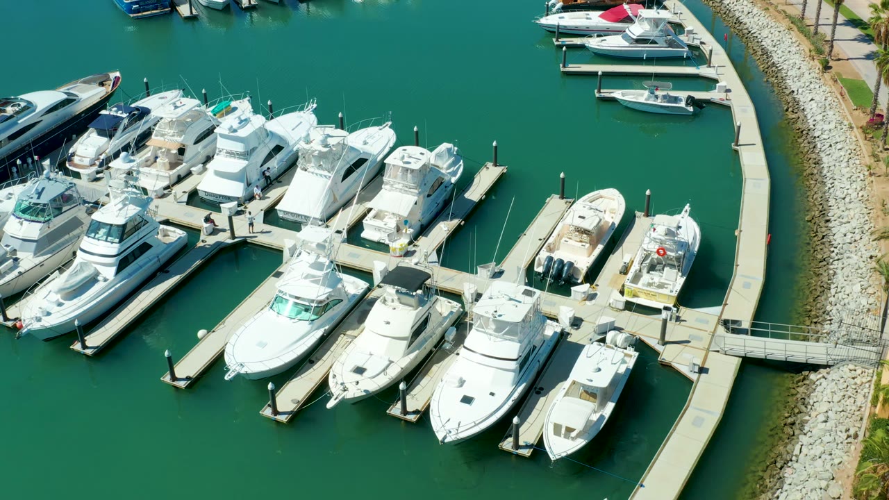 Puerto Los Cabos marina from above, with several white yachts and smaller boats moored to narrow docks, turquoise green water across the frame, shoreline rocks at right, and no visible people.