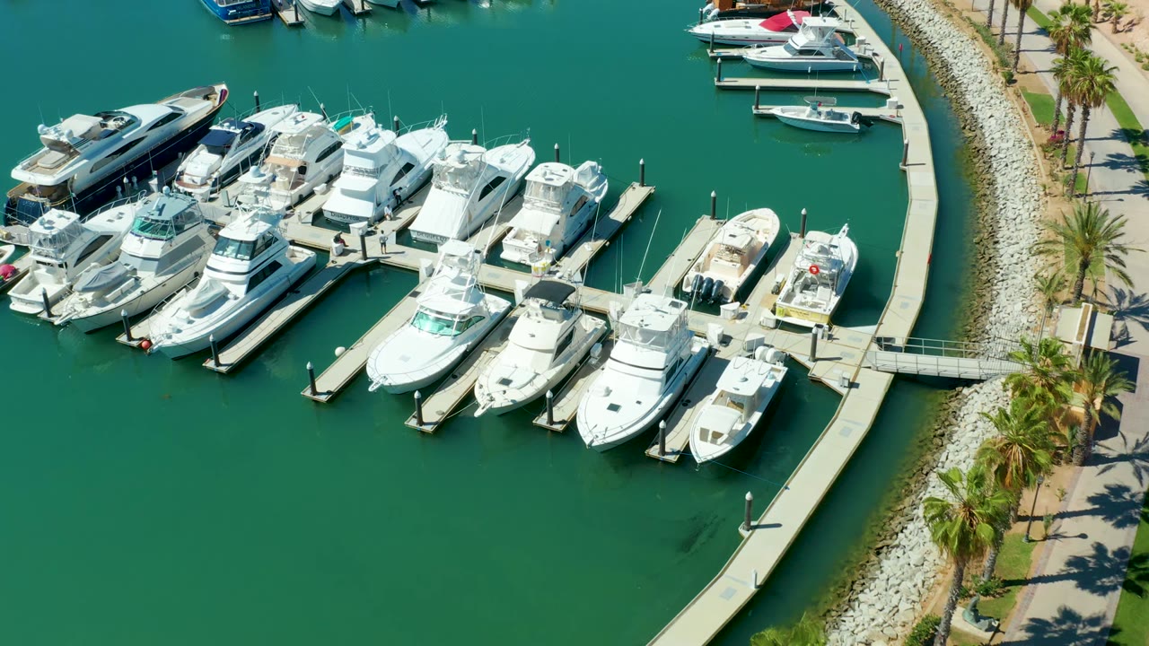 Aerial frame of Puerto Los Cabos marina with white boats along narrow docks, the curved concrete pier crossing the lower right, palm shadows on the shore path, and no visible people.