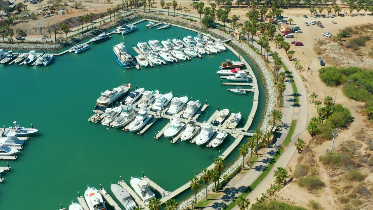 Aerial frame of Puerto Los Cabos marina with white yachts docked in green water, a curved quay, palm-lined path, dry shore, parked cars, and no visible people at the start of the clip.