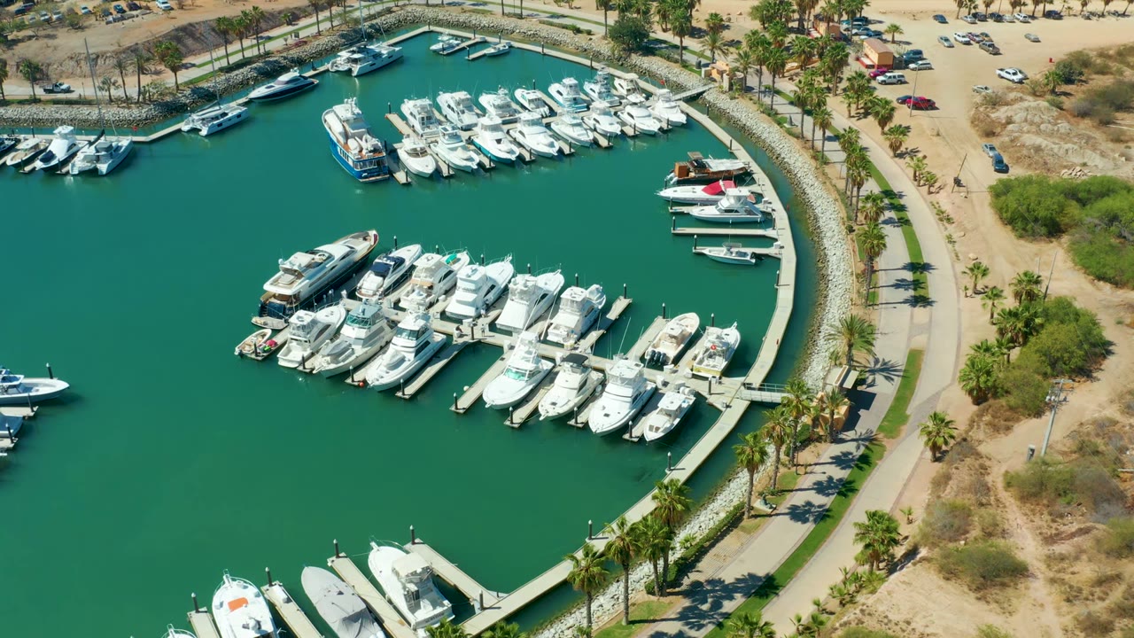 Aerial view over Puerto Los Cabos marina with the main yacht group centered, smaller boats near the upper dock, teal-green water, a palm-lined road edge, dry vegetation, and no visible people.
