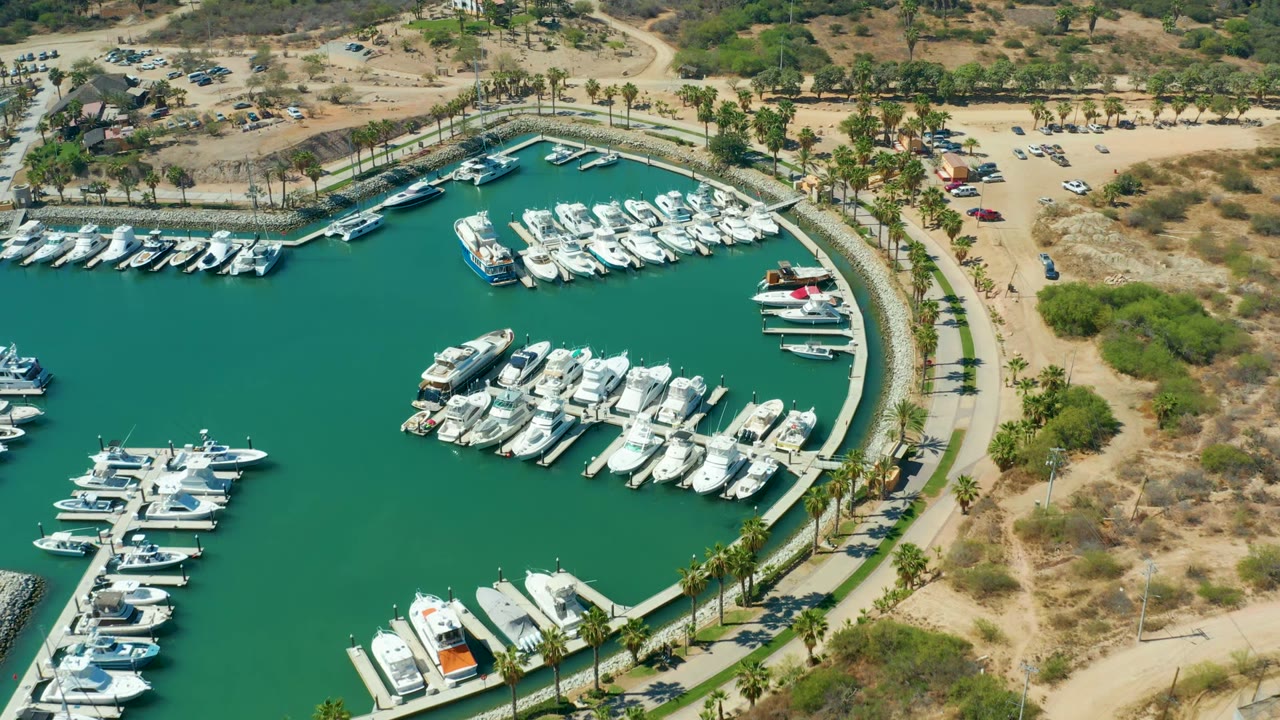 Opening frame of Puerto Los Cabos marina from above, showing green water, clustered white boats, a curved seawall, palm-lined paths, dry sandy ground, parked vehicles, and no visible people.