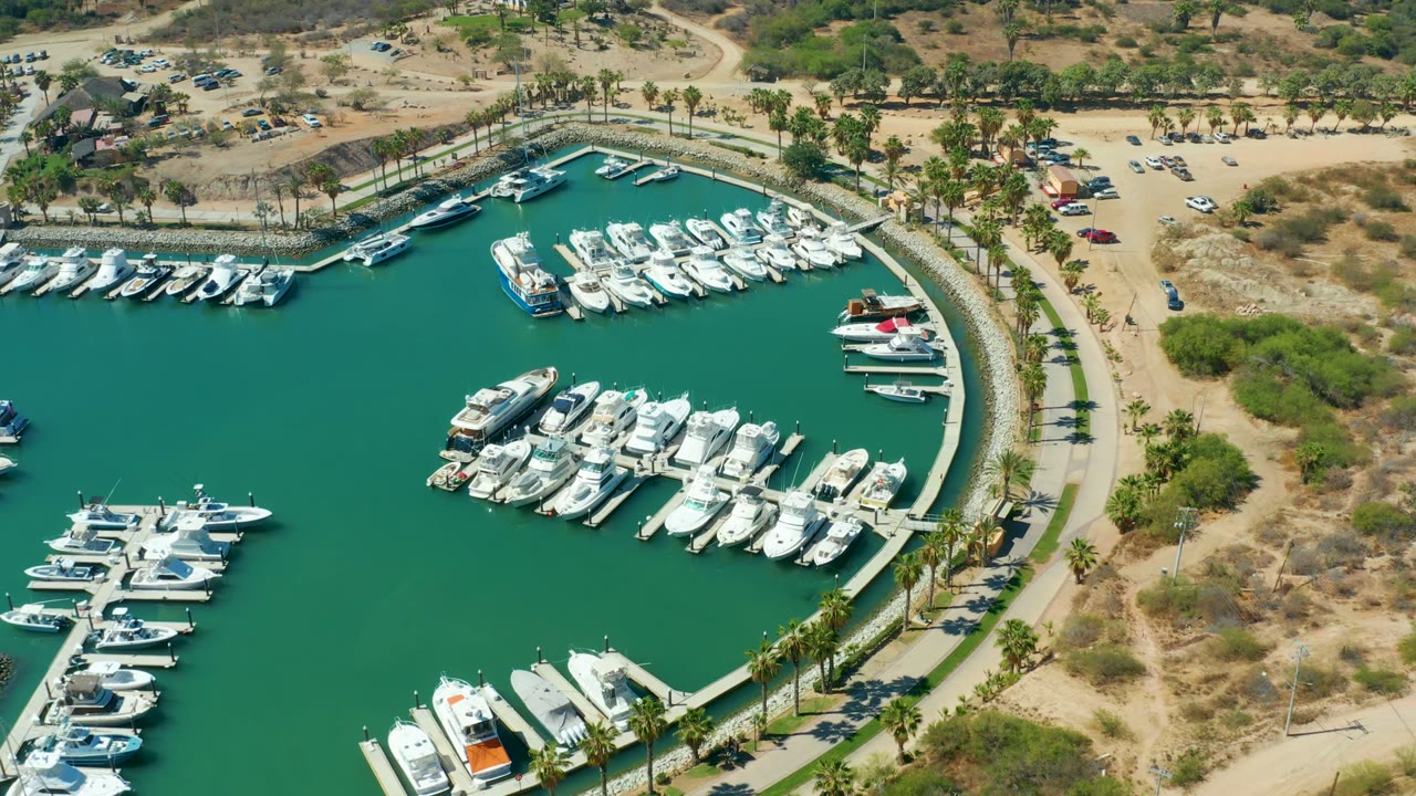 High aerial frame of marina slips with yachts, green water, palms, and sandy parking area.