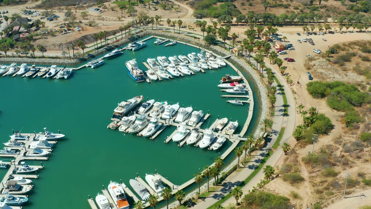At 0:02, the overhead view shows Puerto Los Cabos marina with white boats gathered along the docks, a rounded harbor edge, palms following the path, sandy scrubland, parked vehicles, and no visible people.