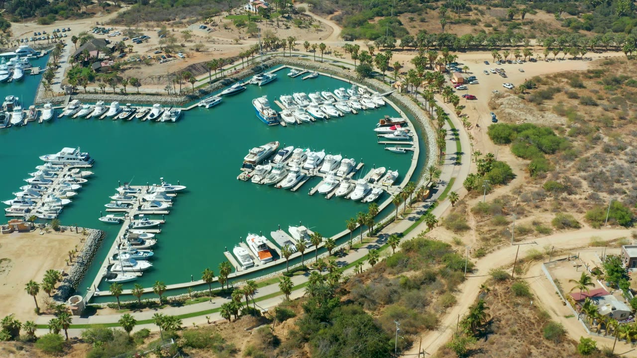 Opening aerial frame over Puerto Los Cabos marina showing turquoise water, clustered white boats, curved docks, a palm-lined road, dry hills, and no visible people.