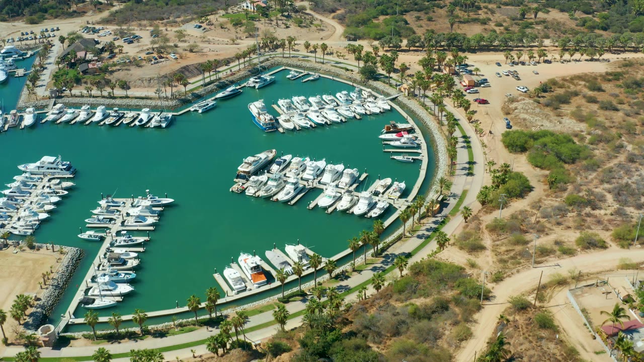 Third frame over Puerto Los Cabos marina showing the curved harbor edge, dense docked boats, palm-lined road, small parking areas, dry scrub hills, and no visible people.