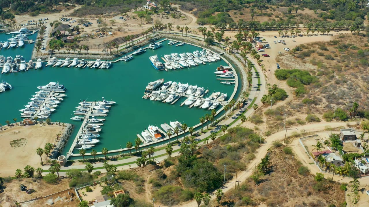 Opening aerial frame over Puerto Los Cabos marina showing white boats in turquoise basins, palm-lined road edges, sandy parking areas, scrub-covered ground, low hills, and no visible people.