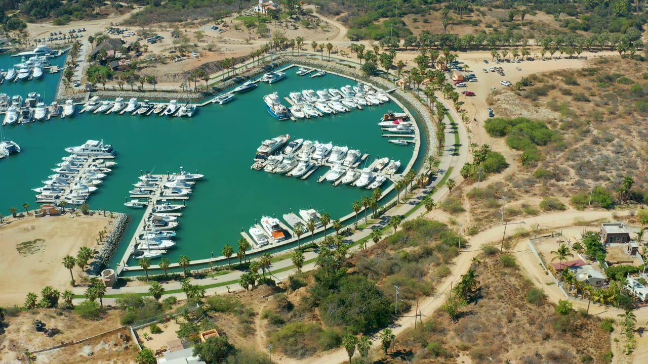 At 0:02, the Puerto Los Cabos aerial shows the marina basin and docked boats slightly farther along the same view, with dry scrubland and parking areas to the right and no visible people.