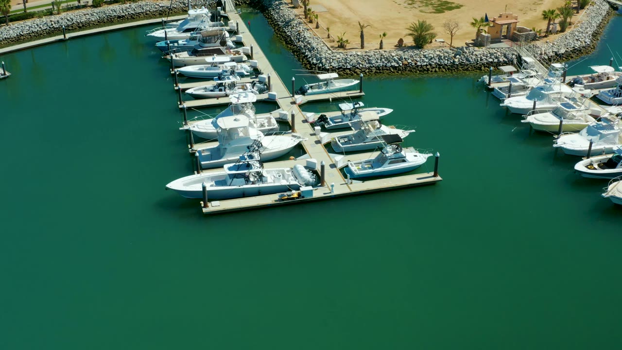Aerial frame of Puerto Los Cabos marina showing docked yachts and smaller boats near the top edge, a sandy shore with palms beyond the rock breakwater, and no visible people.