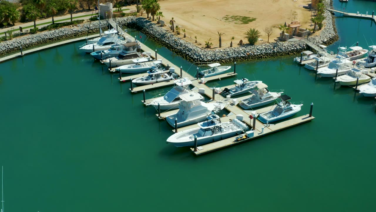 High aerial frame over Puerto Los Cabos marina with white yachts arranged along slips, calm green-blue water filling the foreground, shore rocks and palms above, and no visible people.