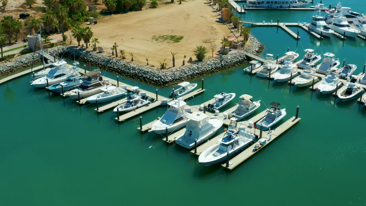 Aerial frame of Puerto Los Cabos marina with white boats tied to floating docks, green water in the foreground, sandy shore, rock breakwater, palms, and no visible people.