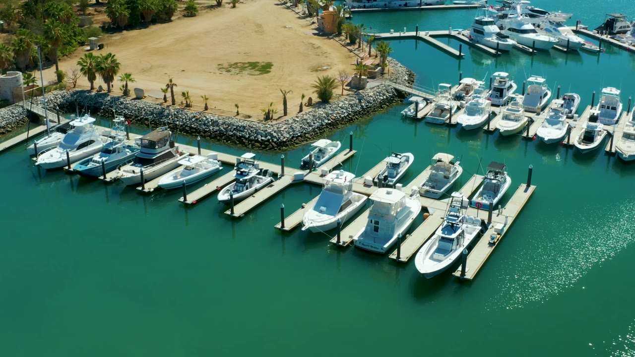 Opening overhead view of Puerto Los Cabos marina with white boats tied to docks, green-blue water, a rocky edge, sandy shore, palms, and no visible people.
