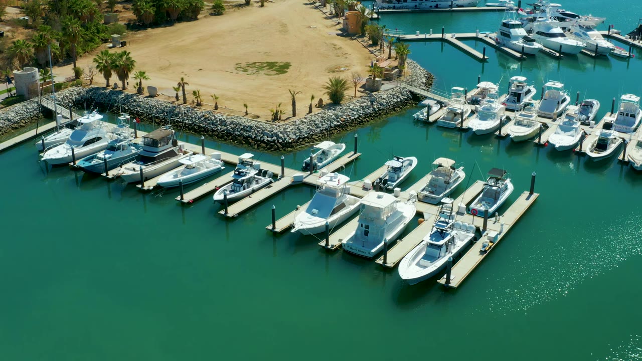 Third frame keeps the Puerto Los Cabos boats and docks in view while the water reflections sit lower in the frame, with sandy ground and palms above and no visible people.