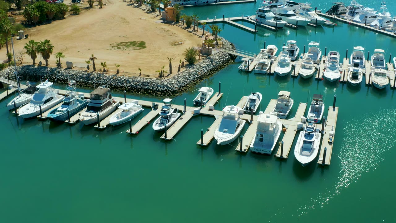 Aerial view over Puerto Los Cabos marina showing white boats tied to docks, turquoise water, sandy shore, palm trees, rock breakwater, and no visible people at the start of the clip.