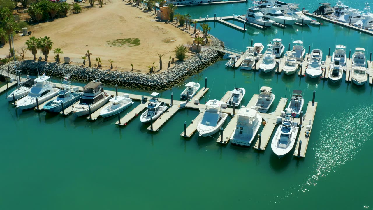 Elevated Puerto Los Cabos marina frame with multiple white boats in slips, green turquoise water, a sandy shoreline, palm trees, a rock breakwater, and no visible people one second in.